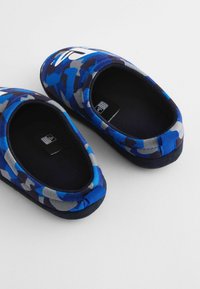 Blå og grå camouflaged slip-on hjemmesko med en blød stofoverdel, rund form og sort fleecefor, prydet med et fremtrædende logo.