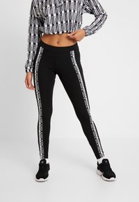 Donna che indossa leggings neri Adidas con strisce bianche con il logo lungo le gambe e un top corto a maniche lunghe abbinato, in piedi con una mano sulla vita.