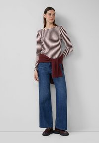 Vrouw die tegen een witte muur staat in een maroon en witte gestreepte lange mouwen top, blauwe jeans, maroon schoenen en een maroon trui om haar taille geknoopt.