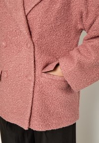 Divkrūšu rozā mētelis, izgatavots no faktūras vilnas, ar pogu aizdari un sānu kabatām. Redzamie šuvumi gar malām.