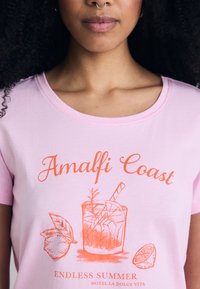 T-shirt de algodão rosa com decote redondo, apresentando um design gráfico laranja de um cocktail e frutas cítricas, rotulado "Costa Amalfitana".
