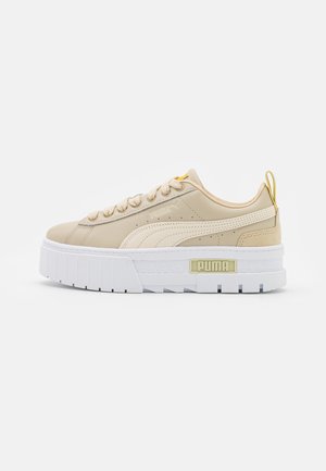 Beige Puma-sneaker med hvid tyk sål, snørelukning foran, beige Puma-stribe og gul hælflig på ensartet hvid baggrund.