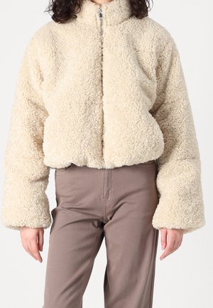 Beżowa kurtka sherpa zapinana na zamek z długimi rękawami, w połączeniu z wysokim stanem taupe spodniami, na osobie na jednolitym tle.