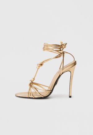 Gold strappy high-heeled sandal with a sleek design, featuring a lace-up ankle strap and knotted detailing on front. Smooth, shiny texture.