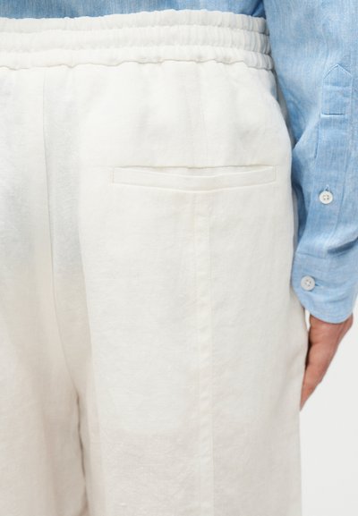 Κοντινή λήψη λευκών παντελονιών με ελαστική μέση και πίσω τσέπη, φορεμένα με μπλε πουκάμισο μακρυμάνικο, εμφανίζοντας το πλάι και το χέρι.