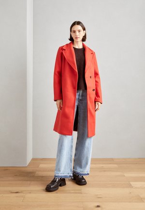 Manteau en laine rouge avec un large col, pull noir en dessous, et jeans larges bleu clair. Des chaussures noires à talons épais complètent le look.