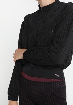 Osoba ubrana w czarną, perforowaną kurtkę na zamek oraz dopasowane spodnie z bordowym pasem w talii i logo Puma na pasie.