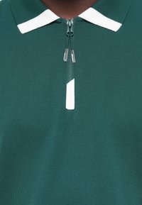 Grøn polo skjorte med lynlås, der har en hvid krave og accenter. Fremstillet af tekstureret stof med en glat finish.