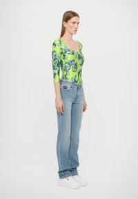 Langermet bodysuit i lys grønn med blå blomster mønster, kombinert med lyseblå jeans og hvite sneakers, som viser en tettsittende design.