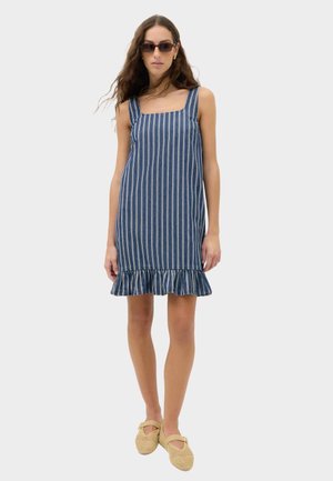 Vrouw met mouwloze blauw-wit verticale gestreepte jurk met gerimpelde zoom, zonnebril, gouden oorringen en beige gevlochten platte schoenen.
