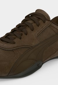Gros plan d'une basket en daim marron avec des lacets assortis, des coutures décoratives et une semelle en caoutchouc foncé sur fond blanc.
