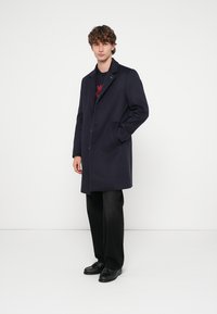 Marineblauwe wollen jas met een getailleerd ontwerp, enkele knoopsluiting en zijzakken, gecombineerd met zwarte broek en schoenen. Gestructureerde stof.