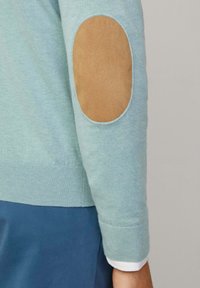 Maglione azzurro chiaro in maglia con toppe in suede marrone ai gomiti, polsini a costine e una texture morbida. Il maglione è abbinato a una camicia bianca a strati.