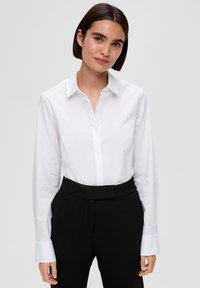 Camicia bianca con colletto e bottoni, maniche lunghe, realizzata in tessuto liscio. Indossata con pantaloni neri aderenti; presenta un design classico.