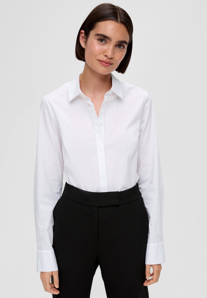 Camicia bianca con colletto e bottoni, maniche lunghe, realizzata in tessuto liscio. Indossata con pantaloni neri aderenti; presenta un design classico.