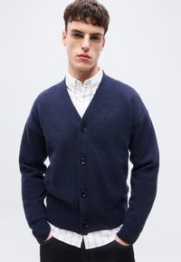 Νέος άνδρας στέκεται με τα χέρια στις τσέπες, φορώντας μπλε σκούρο κουμπωτό ζακέτα πάνω από ένα λευκό ριγέ πουκάμισο, μπροστά σε ένα απλό φόντο.