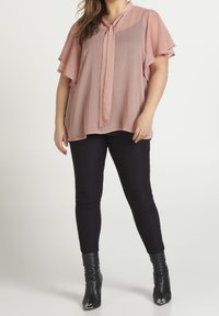 Blouse fluide rose avec des manches courtes à volants et un col à nouer, associée à un pantalon noir ajusté et des bottes à talons noires à bouts pointus.
