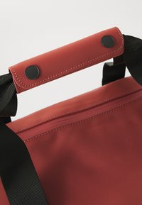 Gros plan sur un tote bag rouge avec un bouton noir et des poignées noires, avec une surface lisse et des détails de couture.