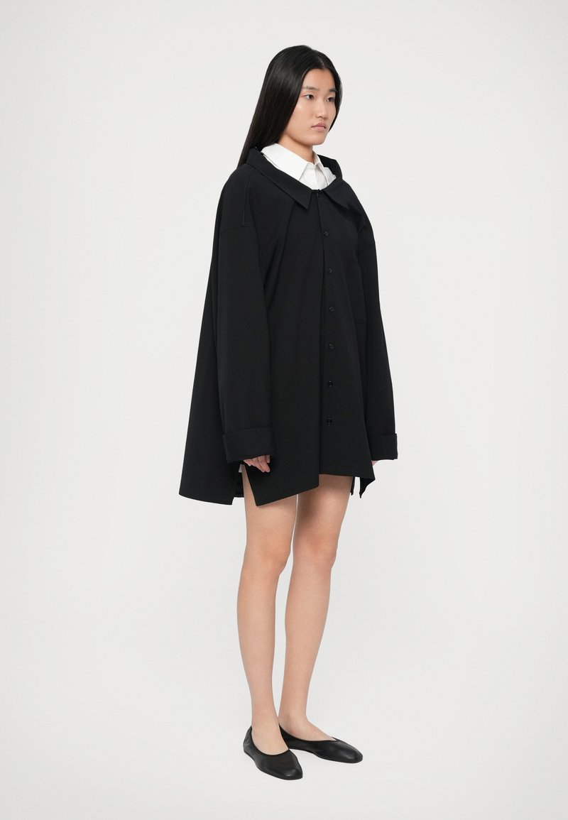 Kvinde iført en oversized sort skjorte med knapper over en hvid skjorte med krave, sorte flade sko, stående mod en ensfarvet lys baggrund.