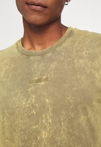 T-shirt vert olive à manches courtes avec une finition texturée, présentant un petit logo brodé au centre, un col rond et un tissu doux.