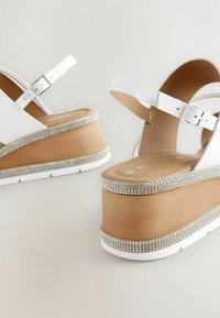 Sandales à plateforme blanches avec une base en bois, dotées d'une bande ornée de strass, d'une sangle de cheville ajustable et d'une texture lisse.