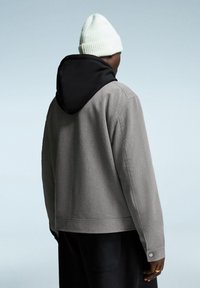 Man wearing a gray jacket with a black hooded inner layer and a white knit beanie, standing against a plain light blue background.