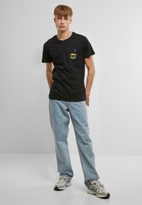 T-shirt en coton noir avec un petit graphique jaune "send nudes", assortie à un jean denim bleu clair et des baskets gris/argent.