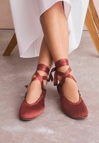 Ballet flats in satin rosso scuro con punta rotonda, caratterizzati da un arricciamento lungo il bordo e lunghe nastri legati attorno alle caviglie.