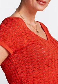 Top de punto rojo con un patrón de olas y hilos metálicos. Presenta un escote en V y mangas cortas, creando un diseño texturizado y transpirable.