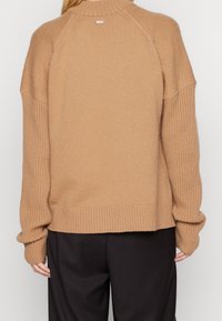 Pull en maille marron avec un col haut, une coupe décontractée, et des poignets et un ourlet côtelés. Présente un petit logo au dos près du col.