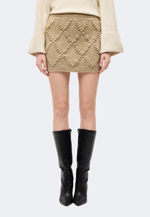 Mini-jupe beige en maille texturée avec motif à losanges, portée avec un pull crème et des bottes en cuir noires montant jusqu'aux genoux.