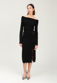 Abito nero scollato sulla spalla con maniche lunghe, spacco laterale, dettaglio della vita a coste e un accento dorato; modella che indossa scarpe con tacco alto decorate.