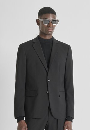 Mann in schwarzem Anzug, schwarzem Rollkragenpullover und übergroßen durchsichtigen Sonnenbrillen, der vor einem einfarbigen hellen Hintergrund steht.