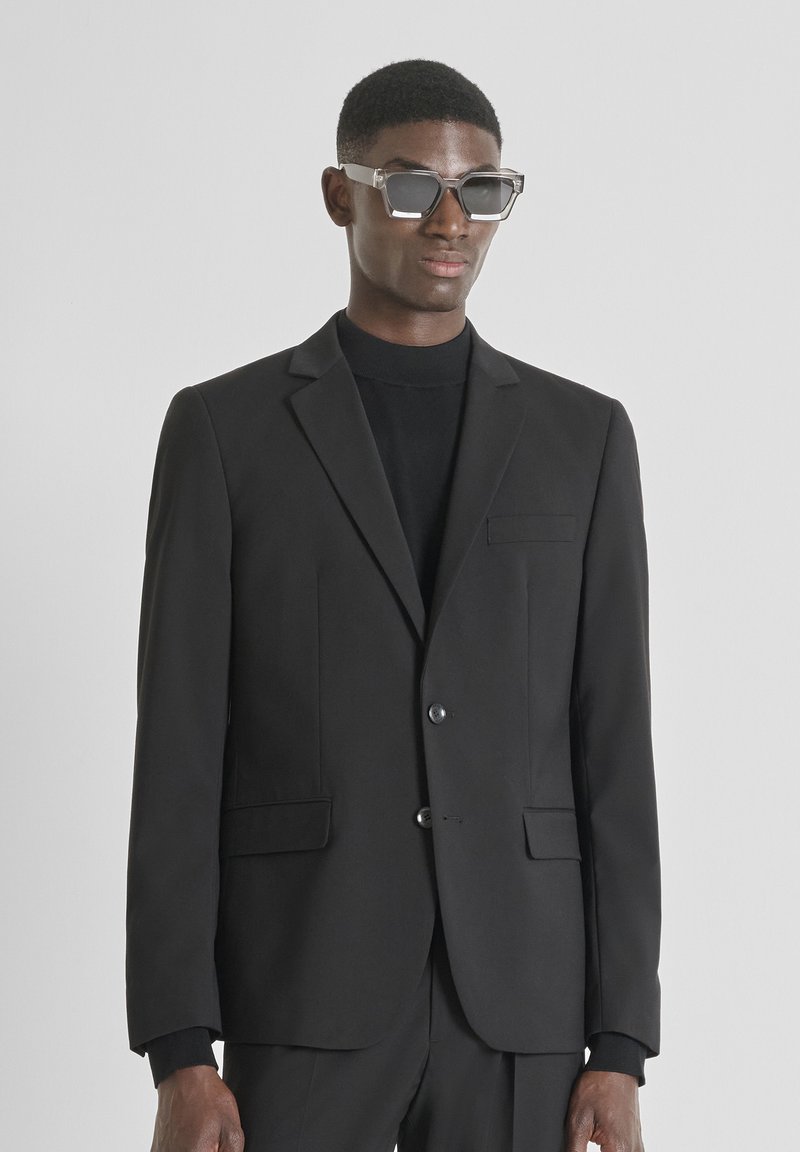 Muškarac u crnom odijelu, crnom dolčeviti i velikim prozirnim okvirima naočala, stoji naslonjen na jednostavnu svijetlu pozadinu.