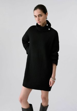 Donna che indossa un abito maglione nero a coste con collo alto abbottonato, orecchini a cerchio in argento, braccialetti e stivali neri fino al ginocchio.