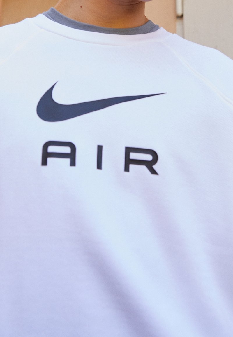 Vit sweatshirt med en svart Nike swoosh-logga och ordet "AIR" tryckt på bröstet, bärs över en grå kragskjorta.