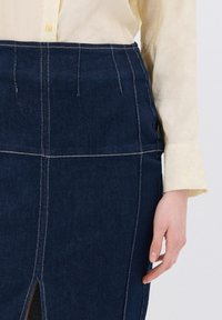 Falda de mezclilla con un tono azul oscuro, que presenta costuras blancas en contraste, una abertura lateral y una cintura estructurada; combinada con una blusa clara.