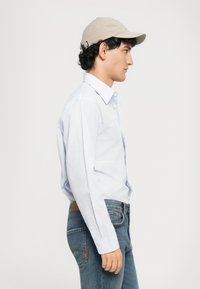 Ung gutt i lyseblå stripete skjorte, blå jeans og beige caps, stående i profil mot en ensfarget hvit bakgrunn.