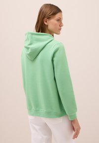 Felpa verde in morbido tessuto con vestibilità rilassata, dotata di cappuccio con lacci, polsini e orlo a costine. Indossata sopra pantaloni bianchi.