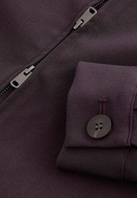 Tamsiai violetinės spalvos marškinėlių audinys su šoniniu užtrauktuku ir rankovėmis, kuriose yra rudas mygtukas. Lygi tekstūra su subtiliomis siuvinėjimo detalėmis.