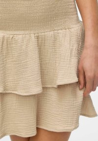 Jupe beige texturée en couches avec un ourlet ondulé portée par une personne, la main reposant sur le côté, montrant les détails et les plis du tissu.