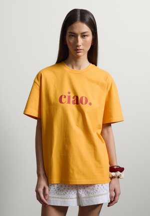 Giovane donna con lunghi capelli scuri indossa una maglietta gialla con "ciao." in rosso e pantaloncini di pizzo bianchi, in piedi davanti a uno sfondo chiaro e uniforme.