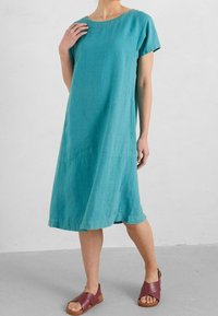Ein türkisfarbenes Leinenkleid mit rundem Ausschnitt, kurzen Ärmeln und lässigem Schnitt, kombiniert mit kastanienbraunen Sandalen mit einem Kreuzriemen-Design.