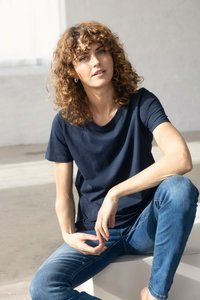 Kobieta o kręconych włosach, ubrana w granatowy T-shirt i niebieskie dżinsy, siedzi w pomieszczeniu, pochylona do przodu z rękami spoczywającymi blisko kolana.