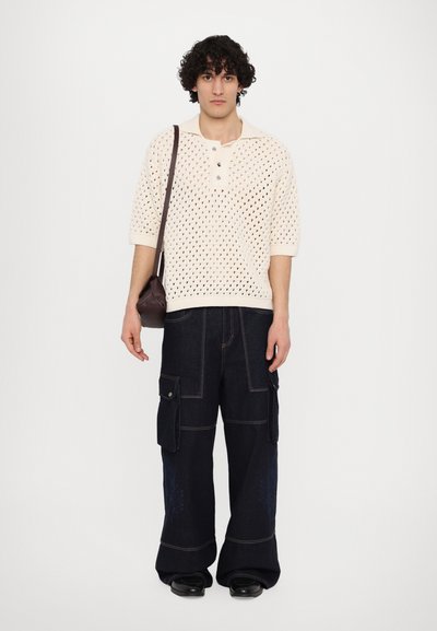 Polo shirt tricoté crème avec un col henley, associé à un pantalon cargo bleu foncé doté de poches latérales et de coutures contrastées. Sac bandoulière marron.