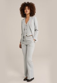 Completo su misura grigio chiaro con giacca aderente, gilet coordinato e pantaloni svasati. Il tessuto liscio e le trame sottili creano un aspetto coeso.