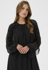 Ung kvinde med langt bølget hår iført en sort gennemsigtig bluse med lange ærmer og flæsedetaljer, stående foran en ensfarvet lys baggrund.