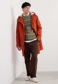 Chaqueta impermeable naranja con capucha texturizada forrada en crema, superpuesta a un suéter verde con patrones, combinada con pantalones marrones y zapatillas blancas.