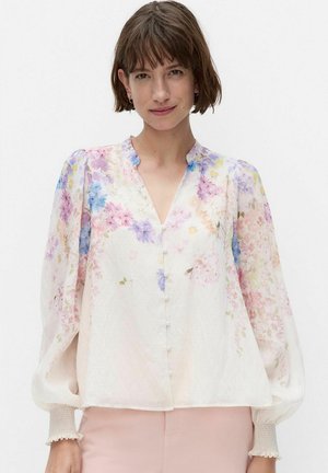 Kobieta z krótkimi brązowymi włosami, ubrana w białą bluzkę z długim rękawem z kolorowym kwiatowym wzorem i perłowymi guzikami, zestawioną z jasnoróżowymi spodniami.