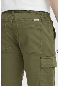 Pantalones cargo verde oliva con cinturilla elástica, múltiples bolsillos y una etiqueta visible. La tela tiene una textura suave y un diseño casual.